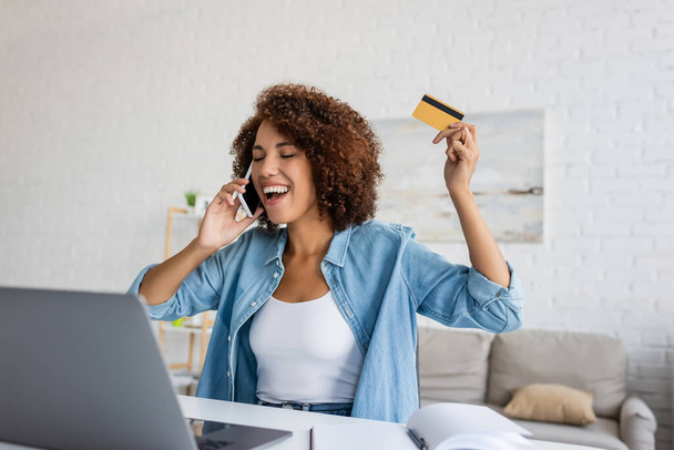 Opgewonden Afrikaans-Amerikaanse vrouw met een creditcard en praten op de smartphone tijdens online winkelen thuis  - Foto, afbeelding