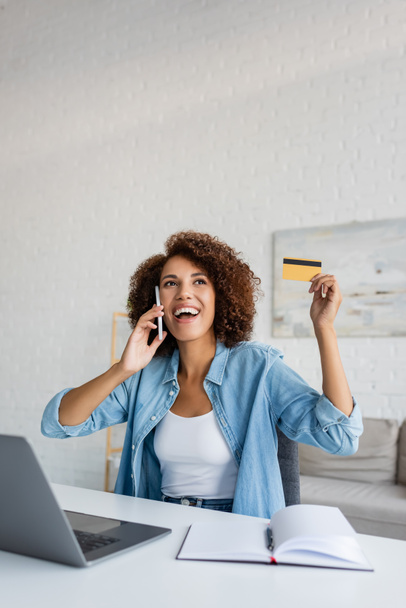 Mujer afroamericana joven sosteniendo tarjeta de crédito y hablando en el teléfono inteligente en casa  - Foto, imagen