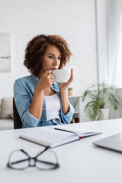curly african american woman drinking coffee and looking at laptop on desk - Photo, Image