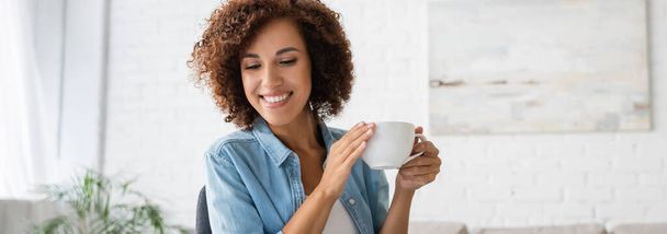 cheerful african american woman holding cup of coffee and looking away at home, banner - Foto, Imagem