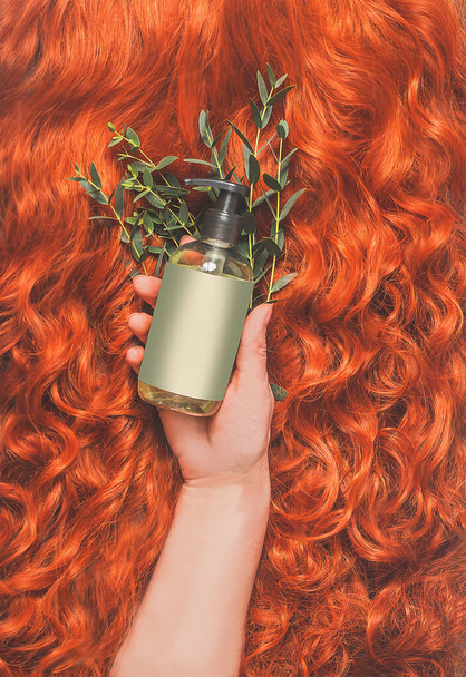 Women hand holding green cosmetic bottle for hair care on curly red hair background with fresh herbs. Top view. - Foto, afbeelding