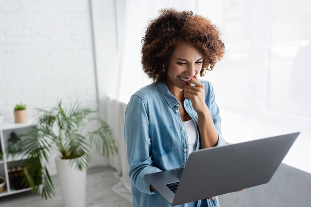 positive african american woman with curly hair holding laptop while working from home - Φωτογραφία, εικόνα