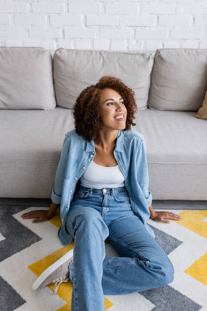 smiling african american woman sitting on rug with pattern near modern sofa in living room - 写真・画像
