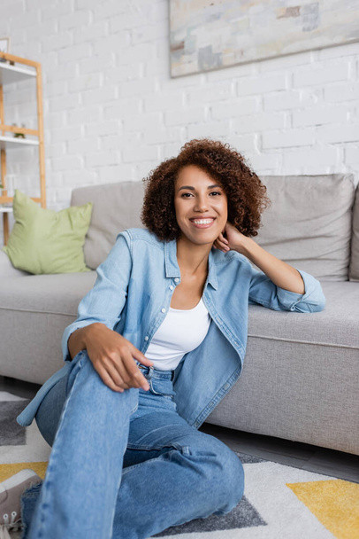 smiling african american woman with curly hair sitting near modern sofa in living room - Fotó, kép