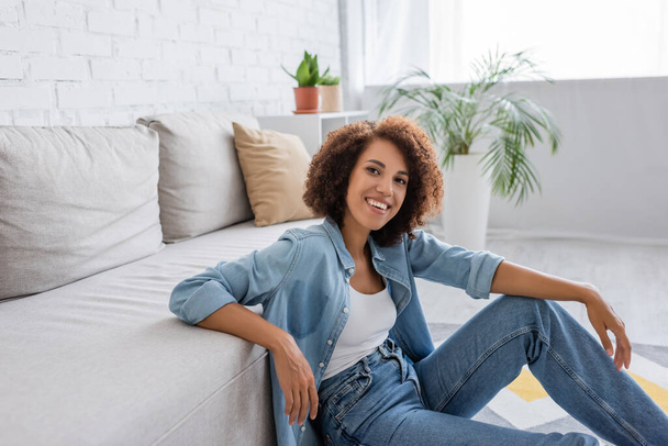 positive african american woman with curly hair sitting near modern sofa in living room - 写真・画像