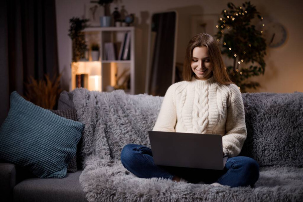Pretty young brunette white sweater sitting on comfortable sofa with crossed legs and typing on wireless laptop. Beautiful smiling woman surfing internet during leisure time at home. - Fotografie, Obrázek