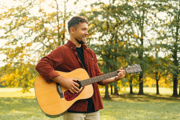 Фото молодого бородатого чоловіка, який грає на акустичній гітарі під час заходу сонця в парку
. - Фото, зображення