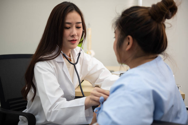 Азійська жінка - кардіолог досліджує жиру жінку, яка уважно слухає серцебиття, використовуючи стетоскоп.. - Фото, зображення