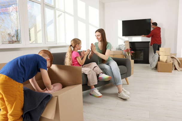 Family settling into new home. Dad setting up TV, happy mom and daughter playing on couch, son unpacking his boxes. Moving day - Foto, imagen