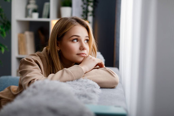 Caucasian lady in casual hoodie lost on her thought while sitting on couch and looking at window. Pensive young woman with blond hair staying alone at cozy home. - Foto, Bild