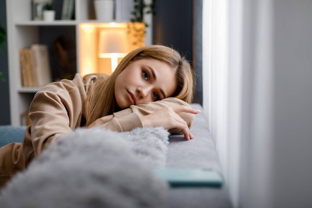 Attractive young woman in casual attire leaning with head on soft grey couch while resting during free time. Daydreaming and relaxation concept. - Photo, Image