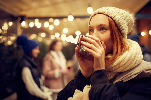 Focus on young smiling beautiful girl in winter clothes drinking hot drinks and dreaming. Spending time with friends at winter fair at evening time. Looks happy, delighted. - Photo, image