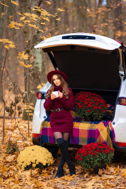 Hermosa chica en acogedor vestido de punto de color burdeos y sombrero sentado en el coche en la naturaleza con fondo otoñal.Bonita modelo con taza de té. Retrato colorido. - Foto, imagen