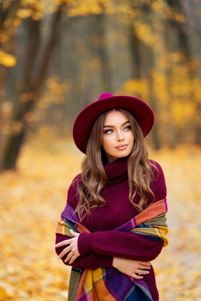 Mooi meisje in gezellig gebreide bordeaux jurk en hoed staande op de natuur met herfst achtergrond. - Foto, afbeelding