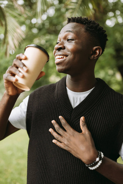 若いアフリカ系アメリカ人の男性の通りの屋外でコーヒーカップに行くに笑みを浮かべて。公園でコーヒーブレイク中の黒人学生。茶色い紙コーヒーカップモックアップ行く - 写真・画像