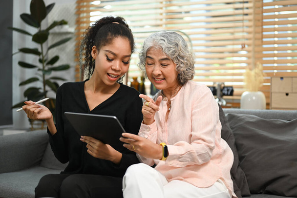 Une jeune femme souriante enseignant à une mère âgée intéressée comment utiliser une tablette ou montre une certaine application. Personnes âgées, concept technologique. - Photo, image