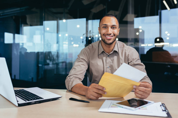 Portrait of happy african american man in office doing paperwork, man looking at camera and smiling holding good news notification bank letter. - Foto, Imagem