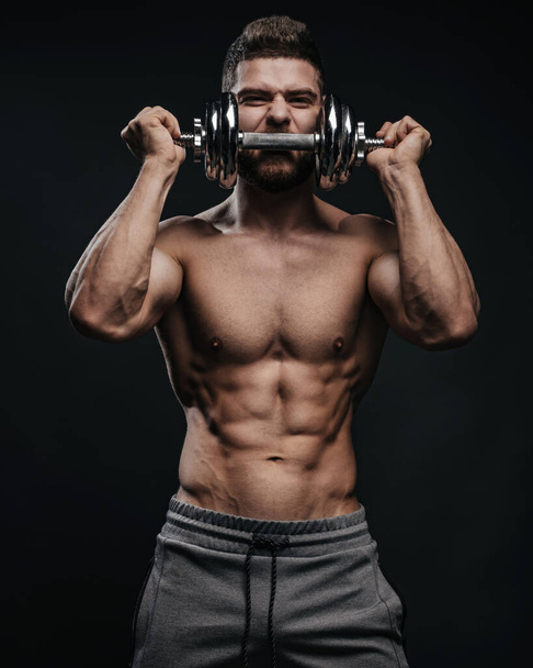 Muskulöser Brocken mit Hantel posiert im Studio. Fitness männliches Modell mit Fitnessgeräten. Hemdloser Mann auf schwarzem Hintergrund. - Foto, Bild