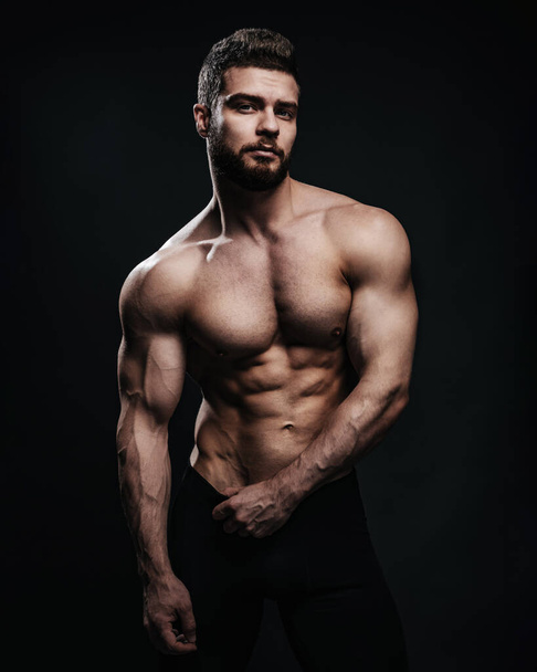 Slim muscular male model at black background. Fitness shirtless guy in black sport pants posing in studio. Man with six pack abs looking at camera. - Fotografie, Obrázek