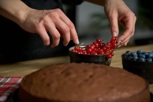 Mulher com uma paixão por cozinhar decora uma fatia de bolo de chocolate artesanal em sua cozinha em casa com amoras e groselhas vermelhas. - Foto, Imagem