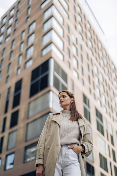 Young pretty woman in cloak walks on street against background of high building. Closeup. - Photo, Image