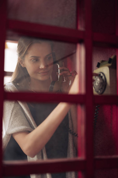 Widok przez szybę do młodej kobiety wybierającej numer telefonu w budce telefonicznej. Zbliżenie. - Zdjęcie, obraz