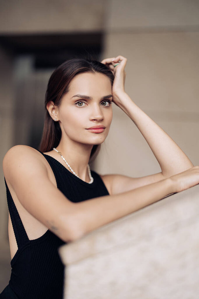 Portrait of young pretty woman in black dress with necklace leaning near building wall. - Photo, image