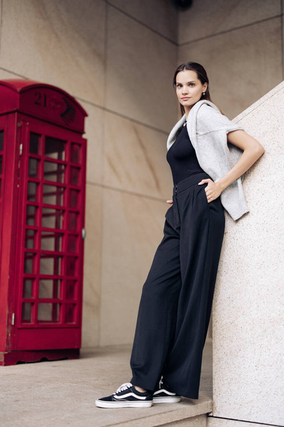 Junge hübsche Frau in schwarzer Hose steht in der Nähe eines Gebäudes vor dem Hintergrund einer Telefonzelle. - Foto, Bild