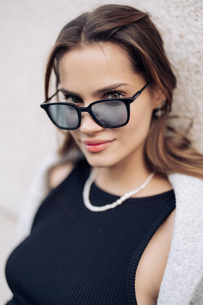 Portrait of young pretty woman in sunglasses and black dress with necklace standing on street near building. - Photo, Image