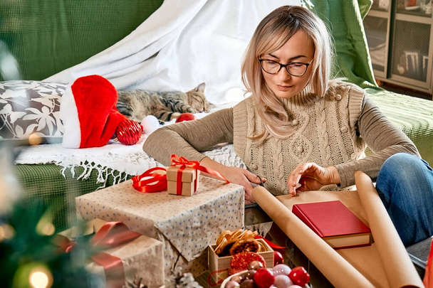 Wrapping christmas gifts. Blond woman wrapping a book as present in recycled card and decorate it with dried oranges and fir branches near the Christmas tree. Winter holiday celebration. - Foto, Bild