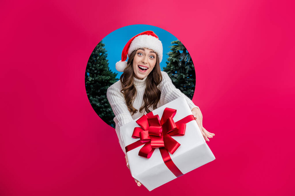 Photo of excited girl dressed santa claus helper x-mas hat giving you gift breakthrough paper hole empty space isolated blue color background. - Photo, image