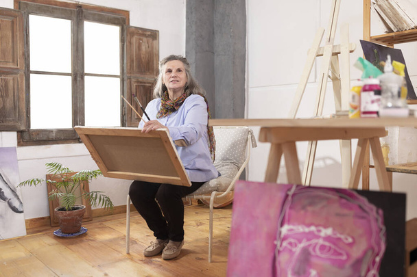 Rijpere kunstenares fantaseert en beeldt zich in hoe ze het schilderij dat ze in haar atelier vasthoudt kan verbeteren - Foto, afbeelding