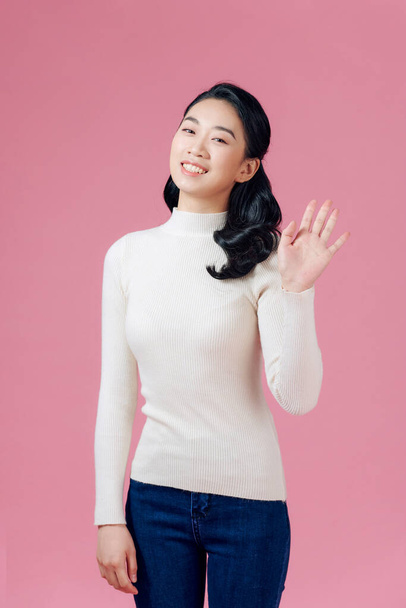 Amistosa, agradable y alegre mujer de Asia Oriental levantando la palma, saludando con la mano agitada y sonriendo sobre fondo rosa - Foto, imagen