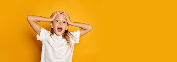 Portrait of little beautiful girl, child in white T-shirt posing with shocked face isolated over yellow studio background. Surprise. Flyer. Concept of childhood, emotions, facial expression, lifestyle - Photo, Image