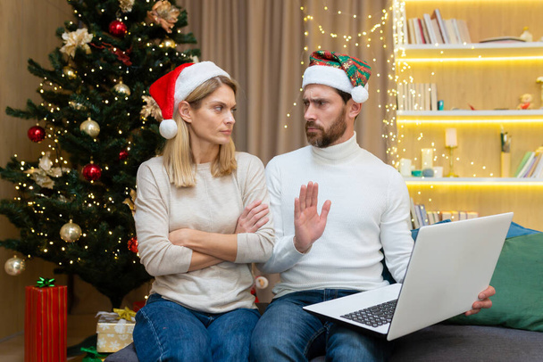 Family conflict on a holiday at homeon Christmas. The woman crossed her arms, jealous of her husband. The man shows his correspondence, conversations on the laptop, shows that everything is fine. - Фото, изображение