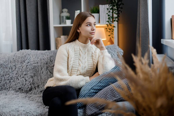 Adorable young woman in casual clothes sitting on comfy couch and looking at window. Caucasian female blonde dreaming alone at home. People and thoughts concept. - Photo, Image