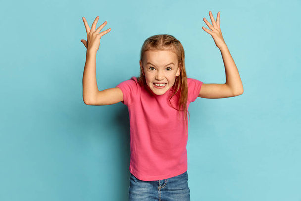 Portrait of little beautiful girl, child in pink T-shirt emotionally posing isolated over blue studio background. Aggressive. Concept of childhood, emotions, facial expression, lifestyle - Zdjęcie, obraz