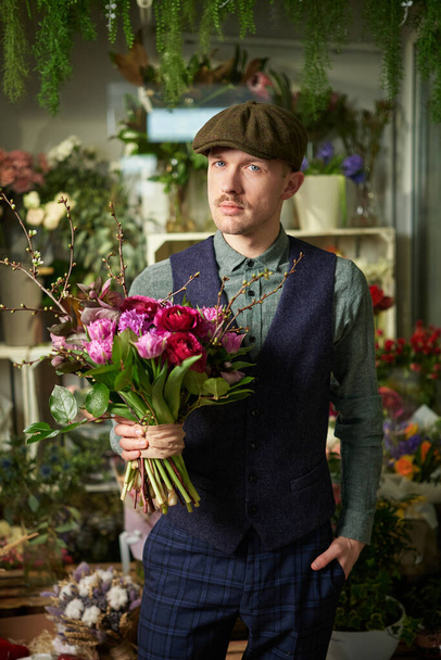 Smiling cheerful caucasian man in peaked cap and old styled clothes from 20s holding beautiful bouquet of red and violet flowers. Mothers Day, Valentines Day or International Womans Day concept - Photo, Image