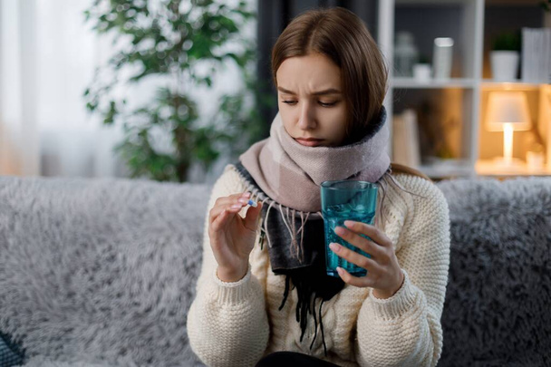 Traurige kaukasische Frau in warmen Schal gehüllt mit einem Glas Wasser und Tabletten, während sie auf der Couch im Wohnzimmer sitzt. Junge blonde Frau nimmt Medikamente gegen Grippeviren. - Foto, Bild