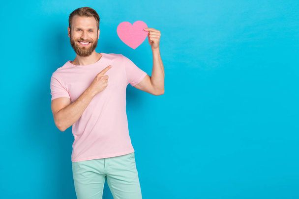 Photo of pretty sweet young man wear pink t-shirt pointing finger paper heart empty space isolated blue color background. - Fotografie, Obrázek