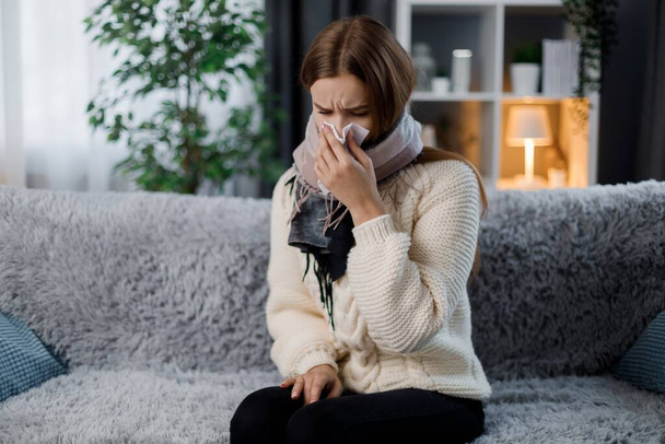 Nezdravě běloška mladá žena v šále fouká nos s kapesníkem, zatímco sedí na pohodlném gauči doma. Koncepce sezónního viru a choroby. - Fotografie, Obrázek