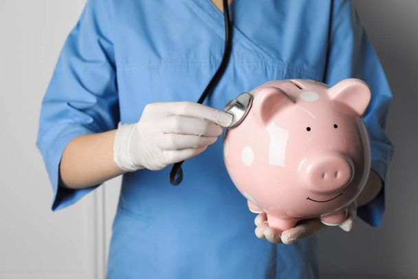 Doctor holding pale pink ceramic piggy bank with stethoscope against white wall, closeup. Medical insurance - Foto, immagini