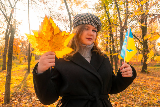 Bosque de otoño. Día soleado. Hojas amarillas cayeron de los árboles. Una niña con un abrigo negro sostiene hojas amarillas y una bandera amarillo-azul de Ucrania en sus manos. Símbolos nacionales de Ucrania - Foto, Imagen