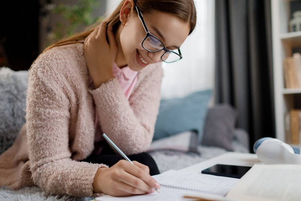 Lächelnde Kaukasierin mit Brille und lässiger Kleidung, die während des Studiums zu Hause in Notizbücher schreibt. Fleißige Studentin verbringt Freizeit für Prüfungsvorbereitung. - Foto, Bild