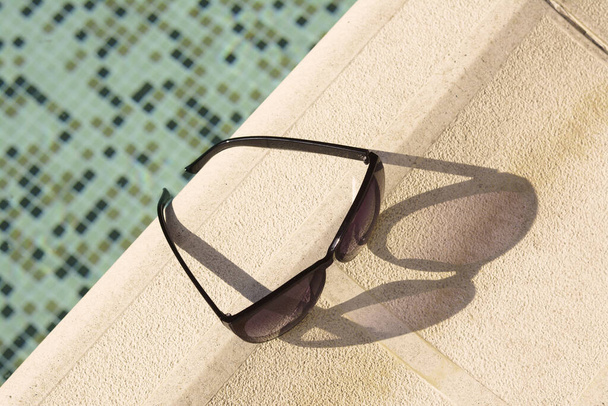 Stylish sunglasses near outdoor swimming pool on sunny day, above view - Фото, изображение