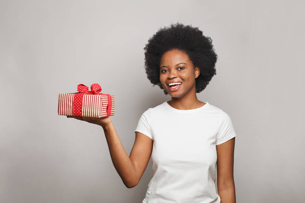 Joyeux jeune femme gaie portant blanc t-shirt souriant et tenant boîte cadeau rouge sur fond blanc bannière murale studio - Photo, image