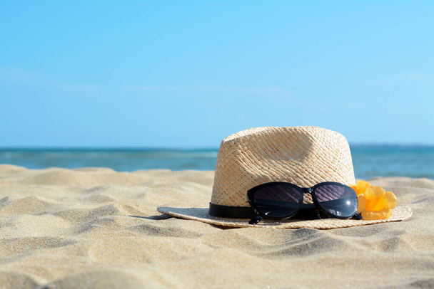 Chapéu com belos óculos de sol e flores na areia perto do mar. Espaço para texto - Foto, Imagem
