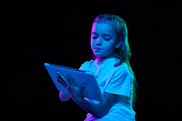 Portrait of little beautiful girl, child in white T-shirt posing with tablet isolated over black studio background in neon light. Concept of childhood, emotions, facial expression, lifestyle - Fotoğraf, Görsel