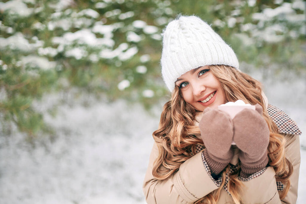 Beautiful blonde girl looks into the camera and smiles. Girl holds a cup of coffee in winter against the backdrop of snow-covered trees. Space for text. New Year's Day, Christmas, St. Nicholas Day. - Фото, изображение