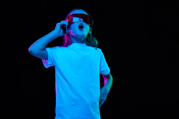 Portrait of little beautiful girl, child in white T-shirt posing in glasses and phone isolated over black studio background in neon light. Concept of childhood, emotions, facial expression, lifestyle - Fotografie, Obrázek
